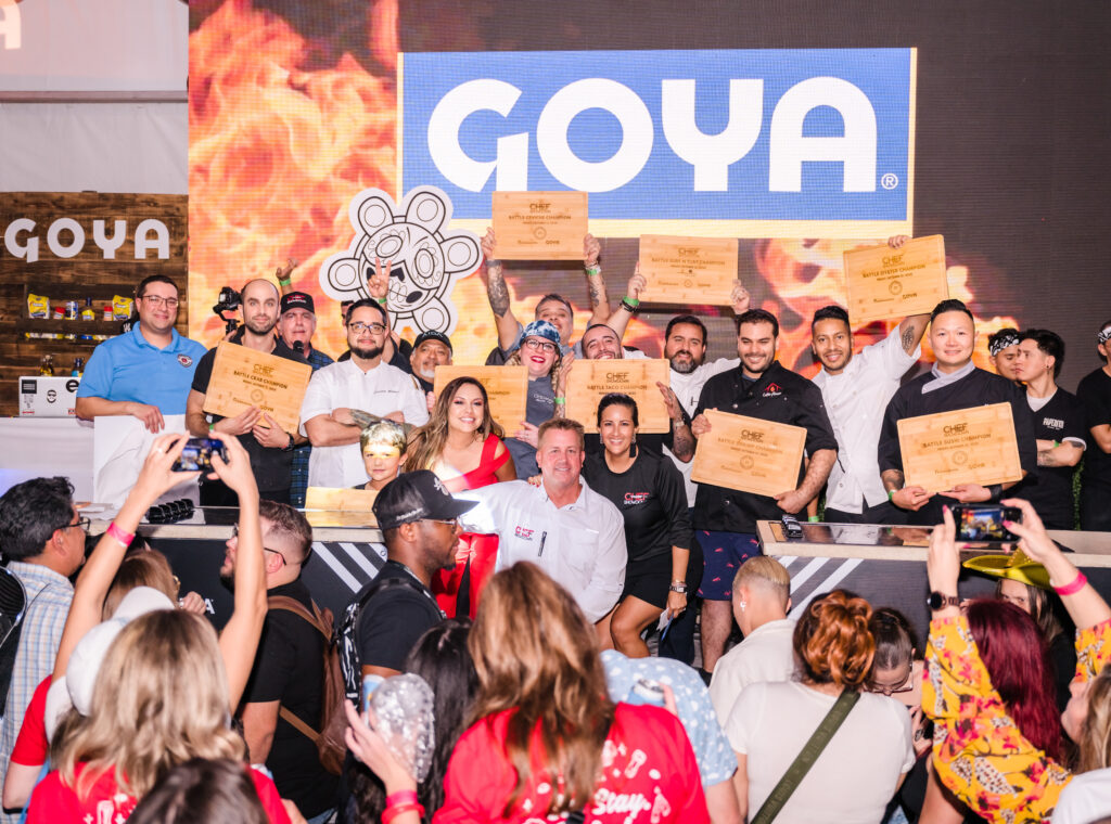 Chefs under a Goya sign, in front of a crowd holding up signs.